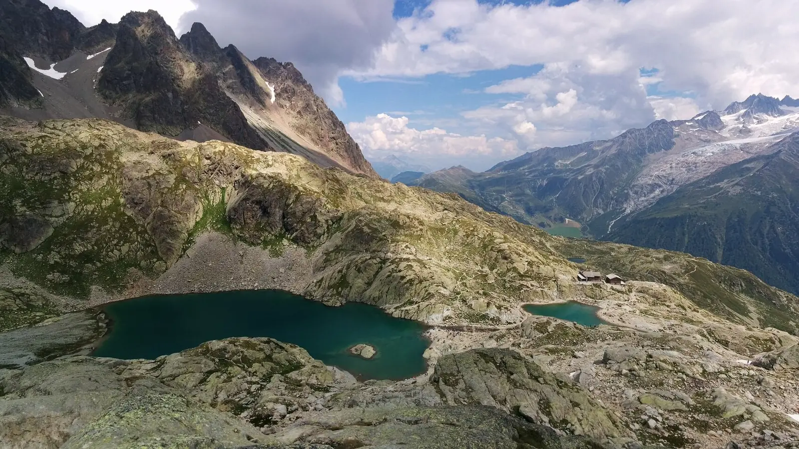 Two turquoise lakes in a bowl of lichen-covered granite slabs with a small refuge beside them, jagged peaks above on the left and glaciers across the valley on the right