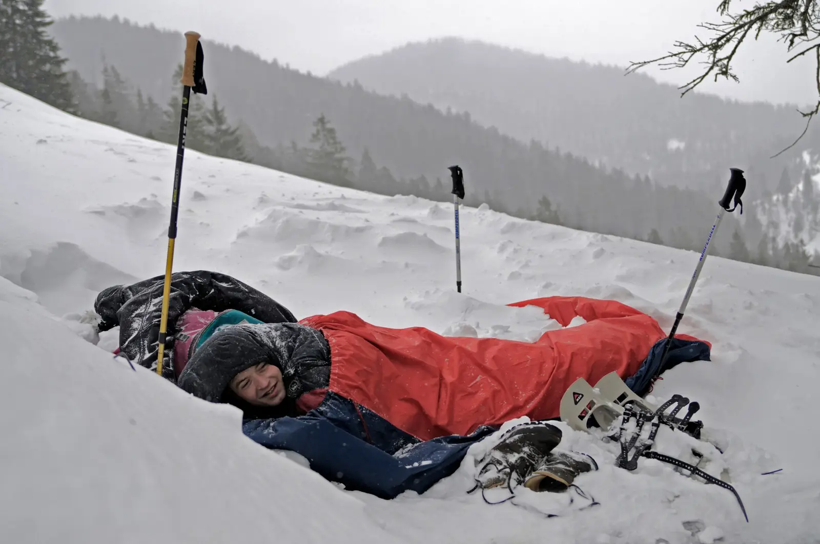 A person lying in a red sleeping bag in a hollow in deep snow, wearing a hooded down jacket, with trekking poles and snowshoes standing in the snow around them