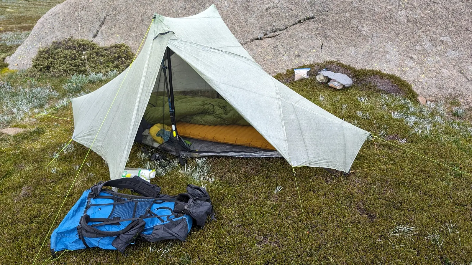 A green sleeping bag lofted on an orange inflatable pad inside an open trekking-pole tent, with a blue backpack lying on the grass outside