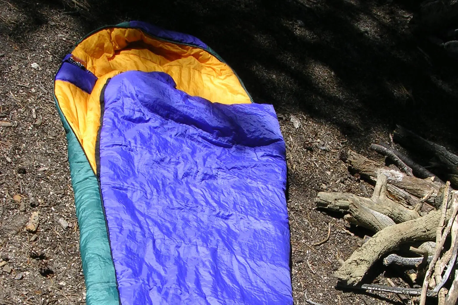 A blue and green mummy sleeping bag lying open on a foam pad on bare ground, its gold-lined hood and neck opening visible at the head end