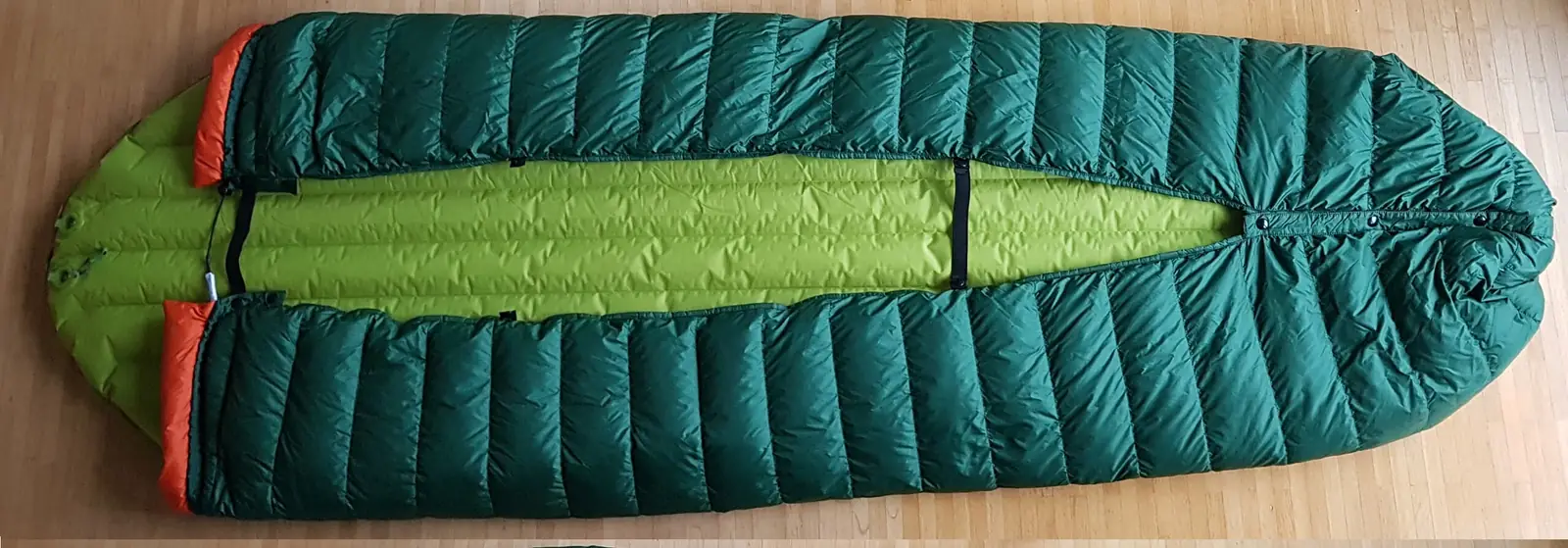 The underside of a green down quilt laid over a sleeping pad on a wooden floor, showing that the quilt is open along the bottom and held around the pad by black elastic straps