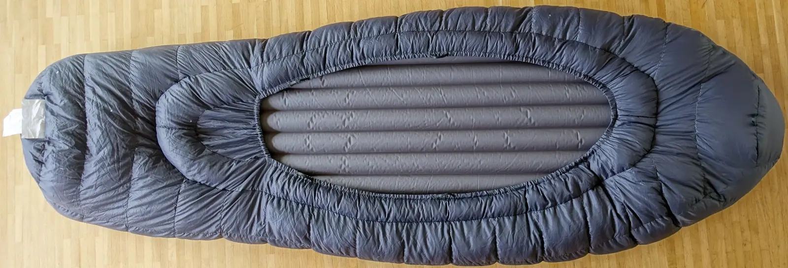 The underside of a gray top-bag sleeping bag seen from above, with a sleeping pad slotted into an oval sleeve cut into the base so no insulation runs beneath it