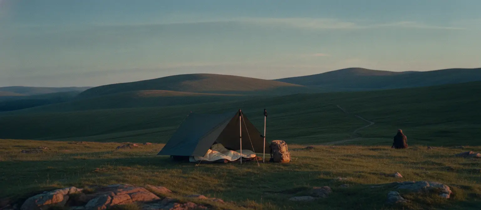 A small A-frame tarp pitched over two trekking poles on open grassland at dusk, with a quilt and a small pack beside it