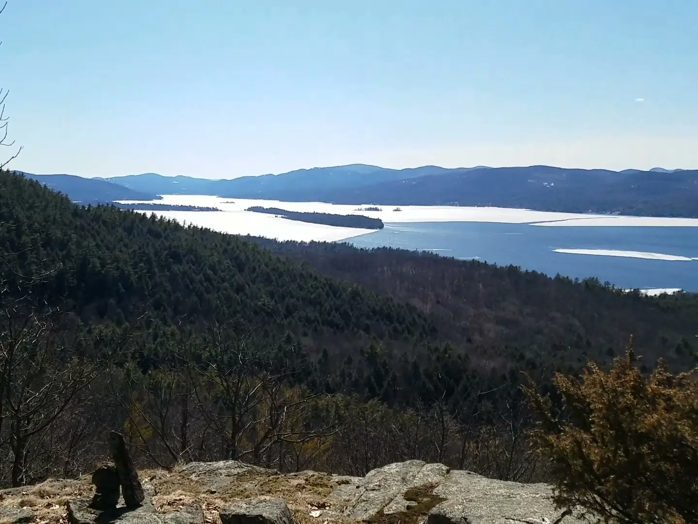 A rocky ledge with bare hardwoods and low brown shrubs in the foreground, above a forested slope falling away to Lake George, where white ice still covers much of the lake below blue mountain ridges