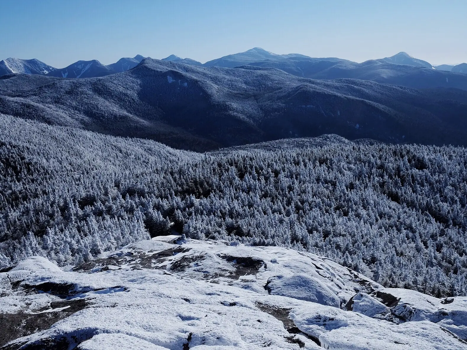 A bare, snow-dusted rock summit in the foreground, above a valley of spruce forest turned completely white by rime, with a line of blue High Peaks on the horizon under a clear sky