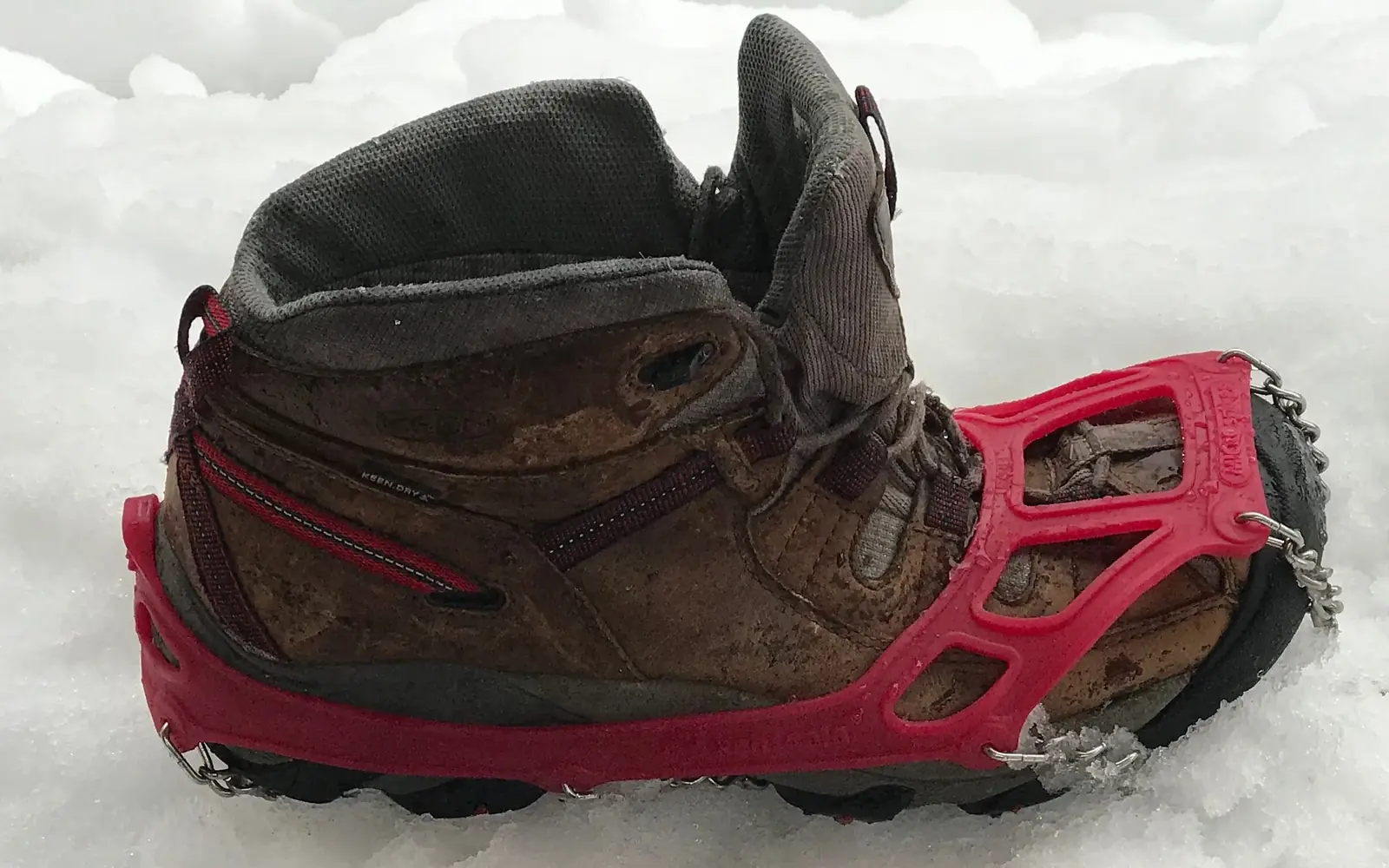 A worn brown leather hiking boot standing in snow with a red rubber traction harness stretched over it, and short chains with small steel spikes wrapped under the sole