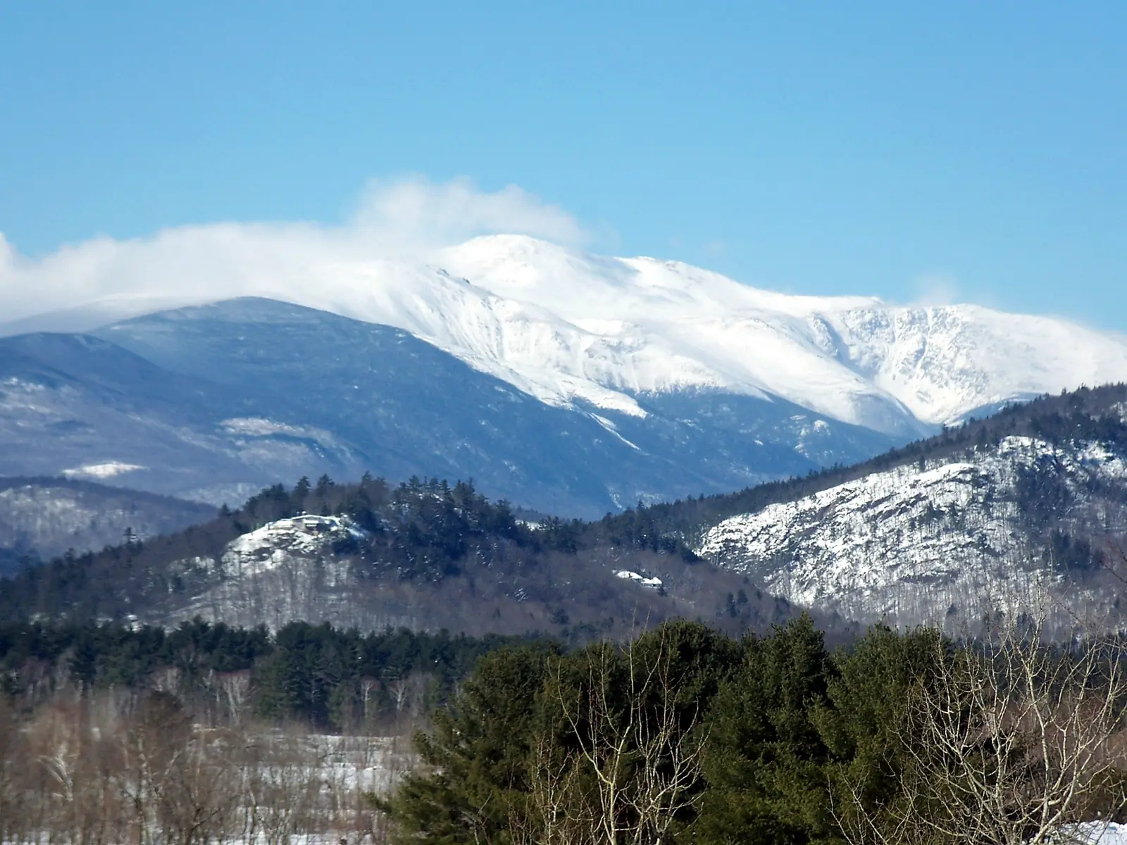 The broad snow-covered summit of Mount Washington under a clear blue sky, with a long plume of snow blowing off the top, above darker forested ridges and a snowy valley