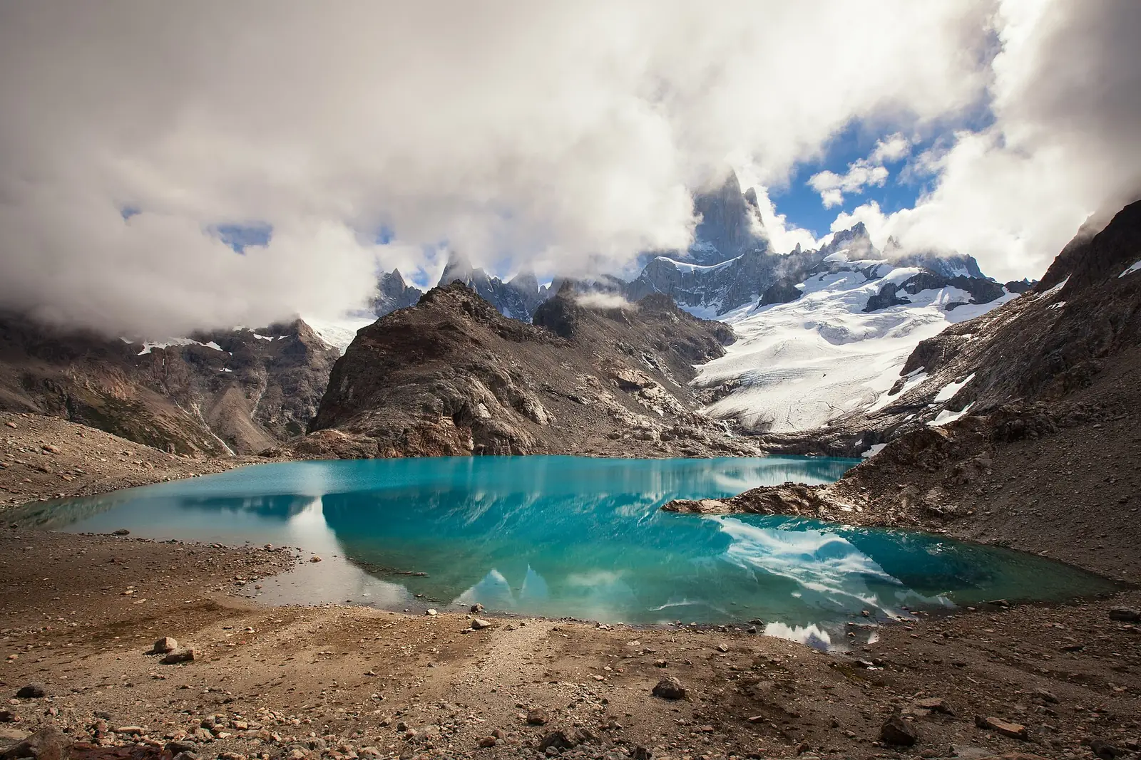 The turquoise Laguna de los Tres with the granite spires of Cerro Fitz Roy rising behind it through broken cloud, glaciers spilling down to the shore