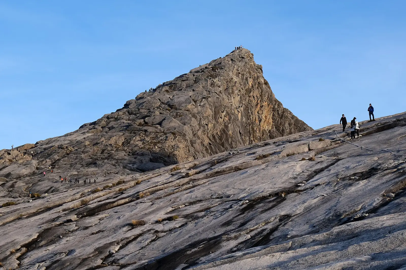 Low's Peak on Mount Kinabalu, a granite fin rising from a vast bare slab of pale rock, with a line of climbers on the summit and a fixed white rope running across the slab