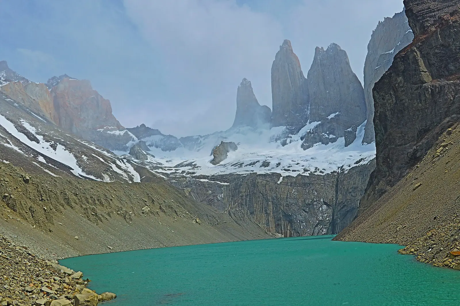 The three granite towers of Paine wreathed in cloud above the green glacial lake at their base, with scree slopes falling to the water on both sides