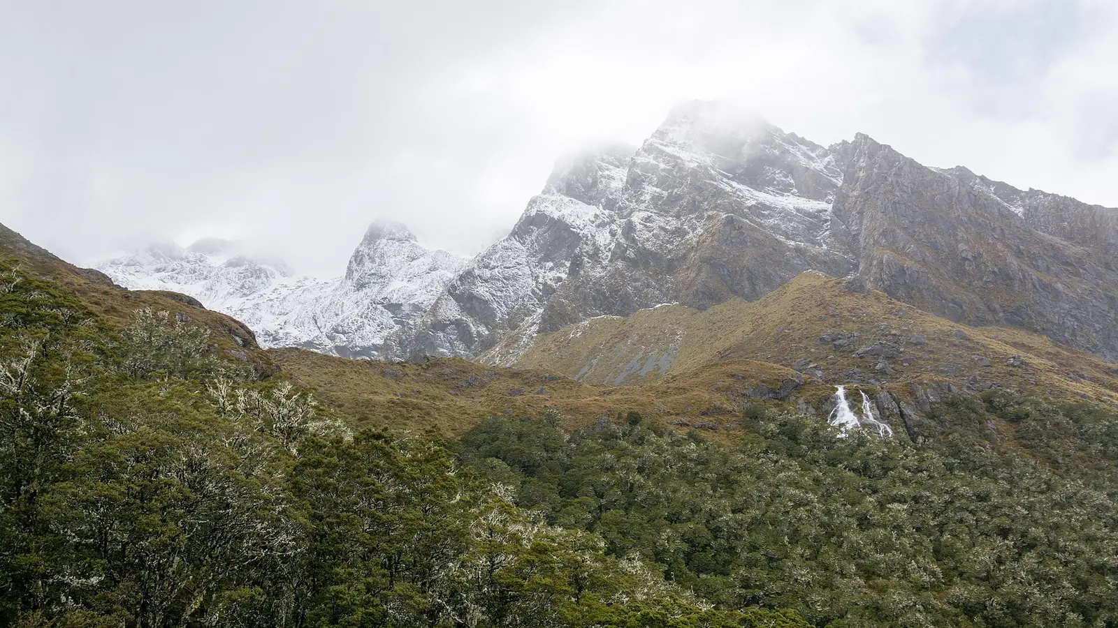 Snow-dusted peaks above beech forest on the Routeburn Track, with tussock slopes, a thin waterfall and low cloud on the summits