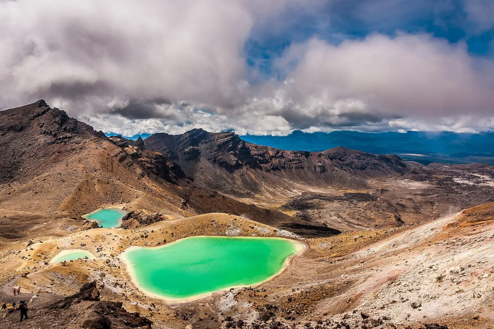 The Emerald Lakes on the Tongariro Alpine Crossing, three vivid green crater lakes ringed by pale sulphur-stained rock, with tiny walkers on the ridge above them
