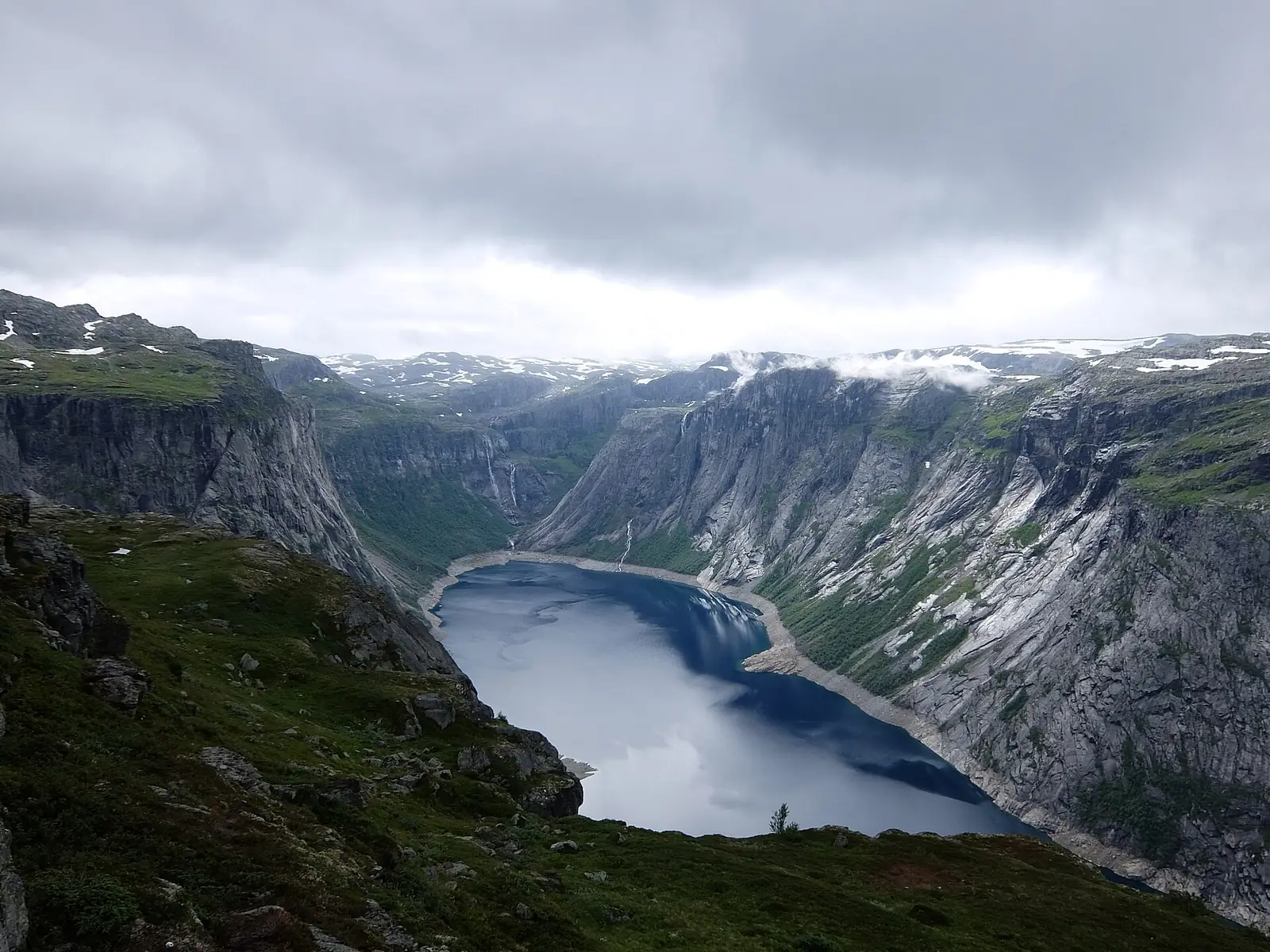 The deep blue lake Ringedalsvatnet seen from high on the Trolltunga trail, hemmed by gray cliffs with snow patches on the plateau above and low cloud over the far end