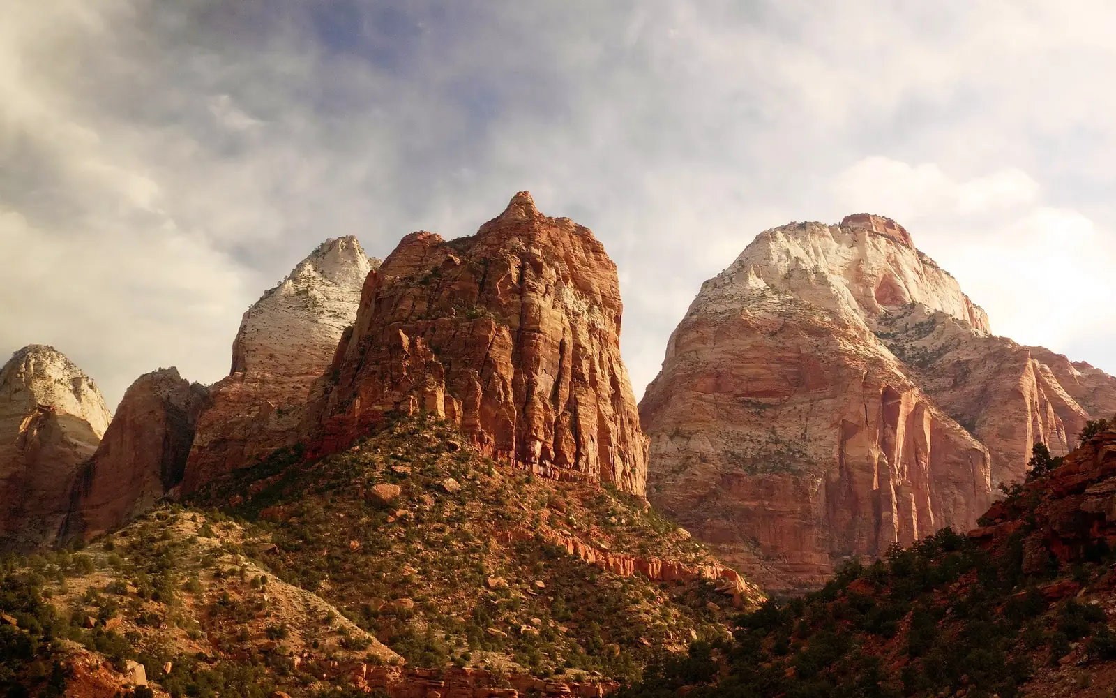 Red and white sandstone peaks in Zion National Park catching low evening light under a bank of cloud, with a wooded ridge below them