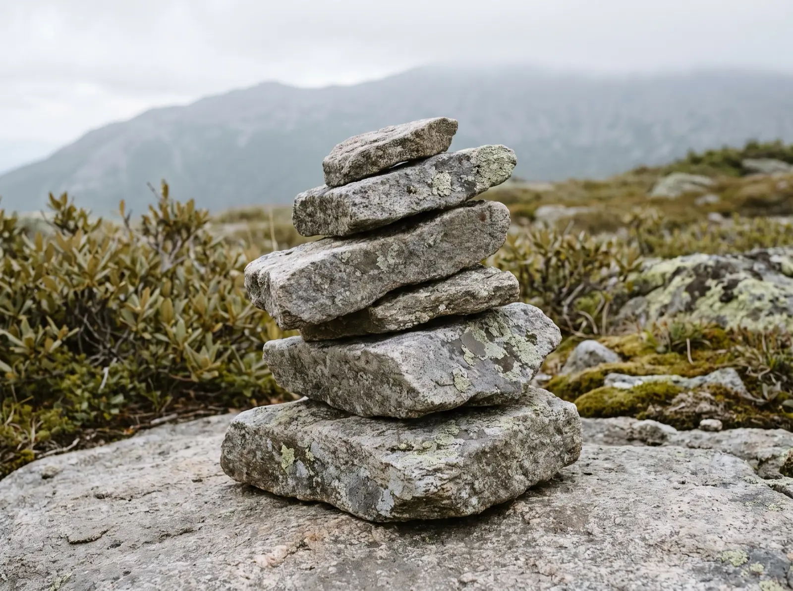 A knee-high stack of five flat lichen-covered granite slabs balanced on bare rock above treeline, with an alpine ridgeline soft behind it.