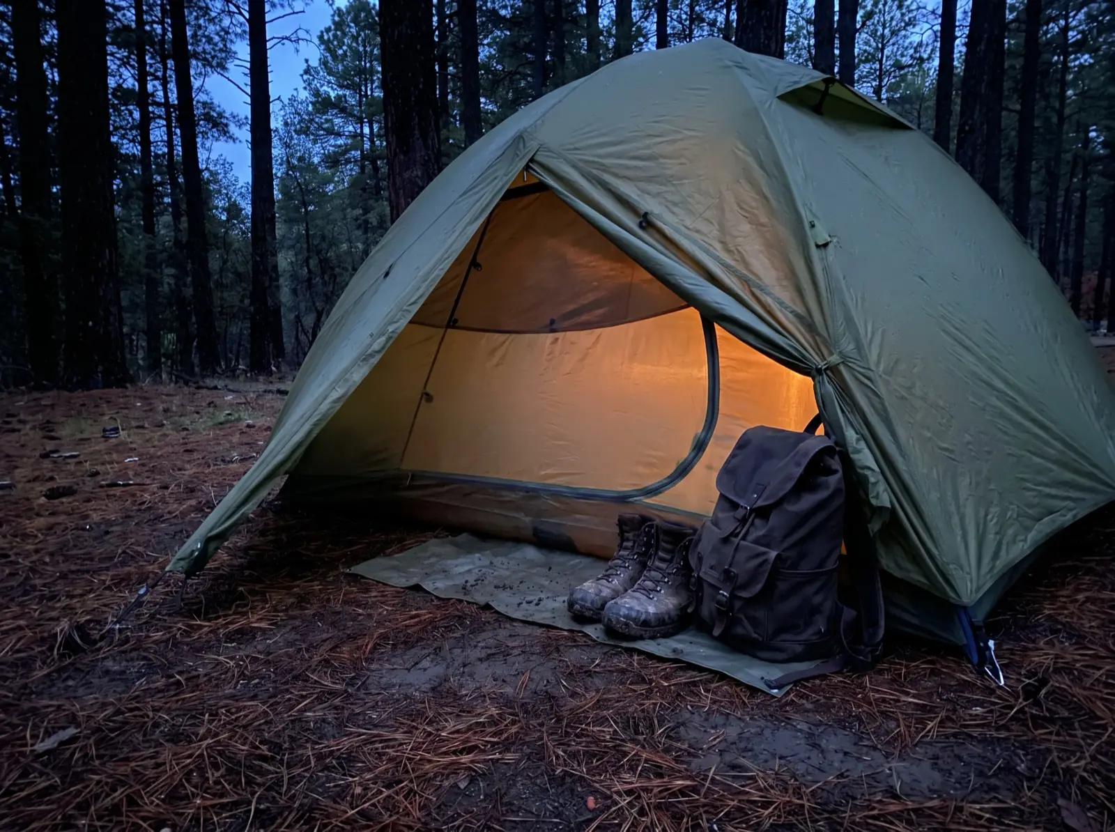 A backpacking tent at dusk with the fly door rolled back, showing muddy boots and a pack stored on a groundsheet in the covered space between the fly and the glowing inner tent.